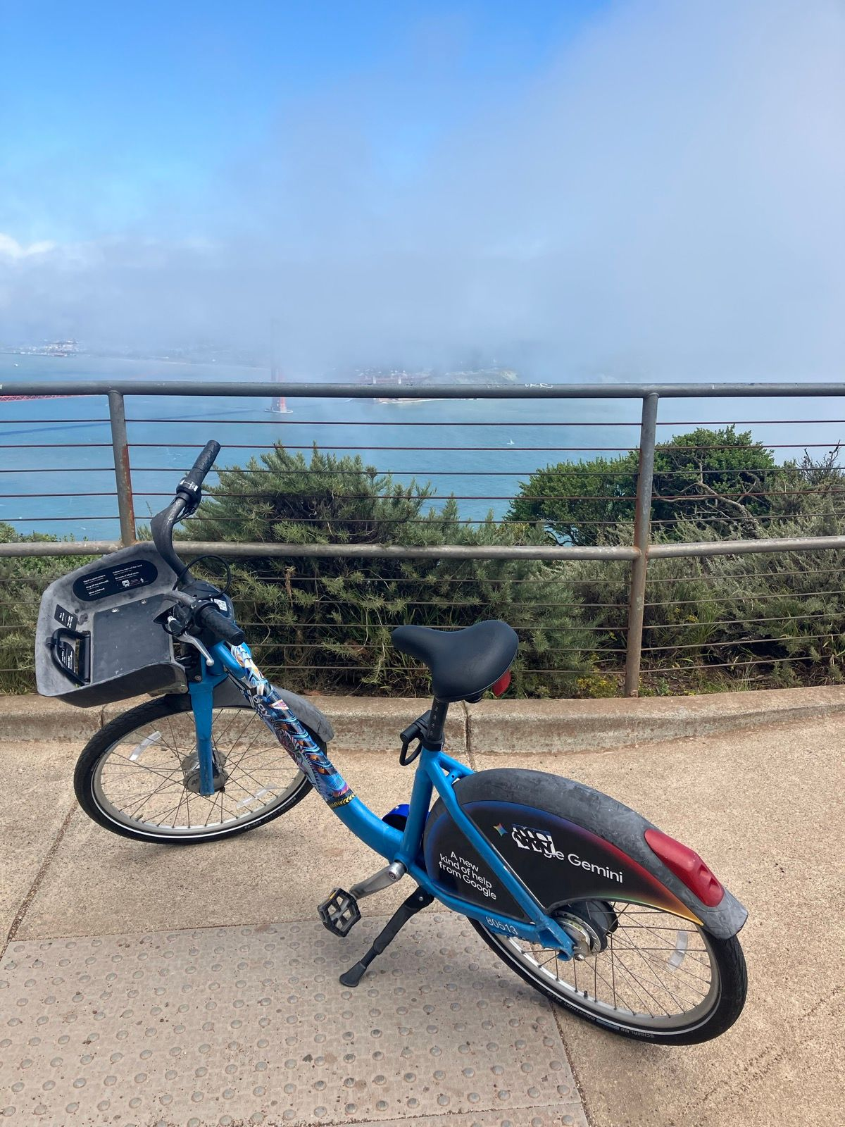A blue bikeshare bike parked on its kickstand overlooking a foggy bay and the towers of a suspension bridge